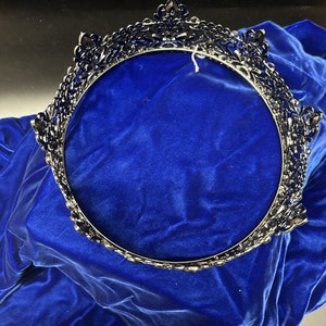 May include: A dark silver-toned crown with ornate detailing, set against a vibrant blue velvet backdrop. The crown features a circular design with intricate patterns and embellishments, suitable for special occasions.