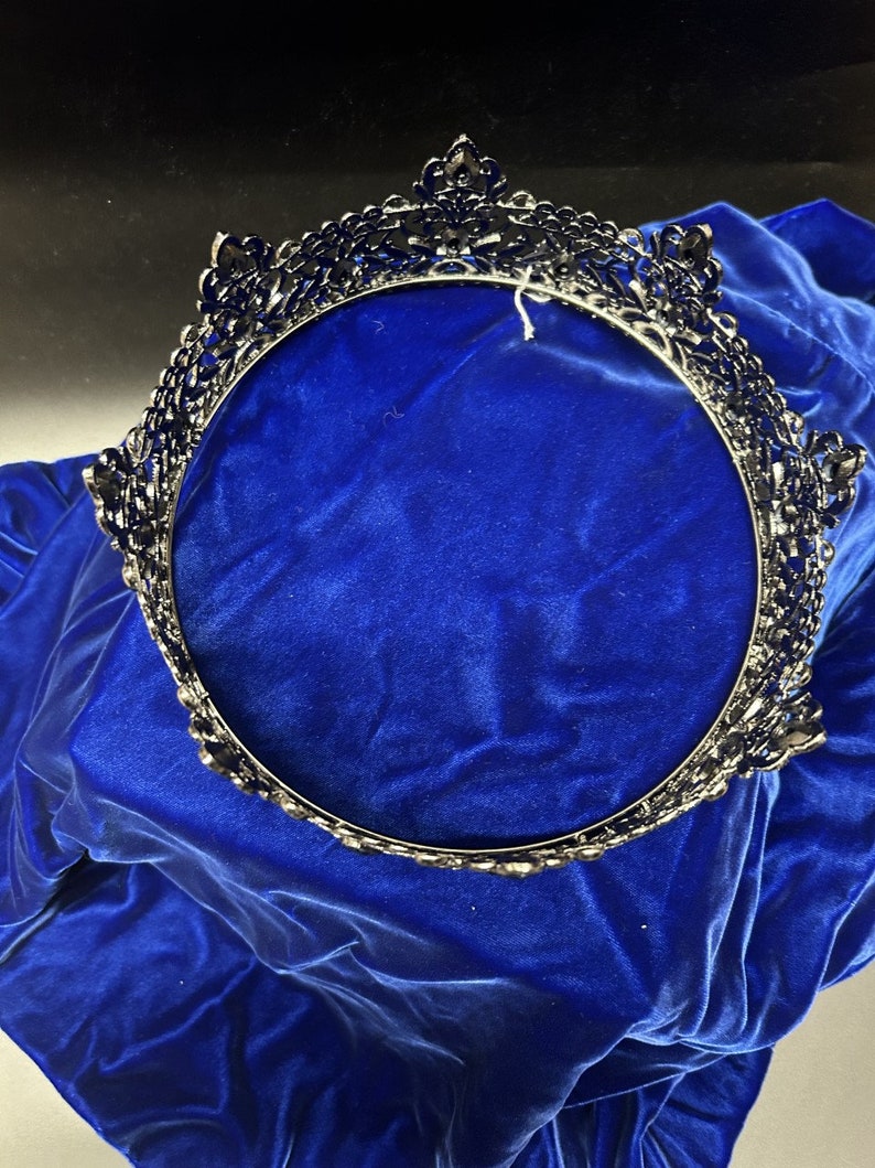 May include: A dark silver-toned crown with ornate detailing, set against a vibrant blue velvet backdrop. The crown features a circular design with intricate patterns and embellishments, suitable for special occasions.