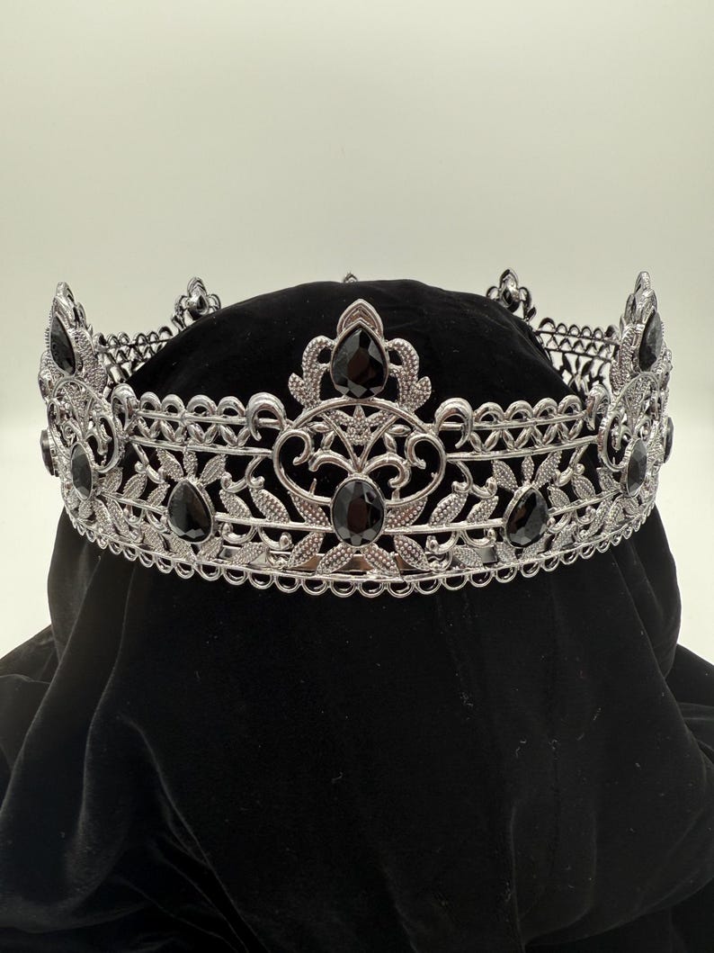 May include: A silver-toned crown with intricate filigree and leaf designs. The crown is adorned with several large, black, teardrop-shaped gemstones. The crown is displayed on a black velvet surface, creating a striking contrast.