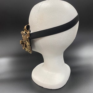 Peut inclure: Un masque de style steampunk avec des engrenages et des accents en laiton, attach&eacute; &agrave; un &eacute;lastique noir. Le masque est pr&eacute;sent&eacute; sur une t&ecirc;te de mannequin blanche.