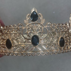 May include: A gold-colored crown with intricate filigree detailing. The crown features three large, black, oval-shaped gemstones and two teardrop-shaped gemstones. The design includes floral and leaf patterns, creating an elegant and regal appearance.