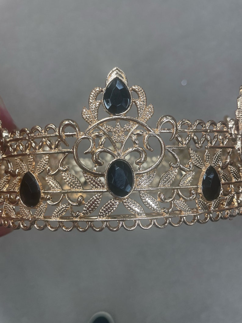 May include: A gold-colored crown with intricate filigree detailing. The crown features three large, black, oval-shaped gemstones and two teardrop-shaped gemstones. The design includes floral and leaf patterns, creating an elegant and regal appearance.