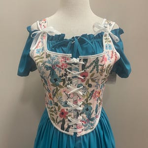 May include: A teal-colored dress with a floral patterned corset. The corset features a white base with a colorful floral design, white trim, and white lacing. The dress has short sleeves and a tiered skirt, displayed on a mannequin.