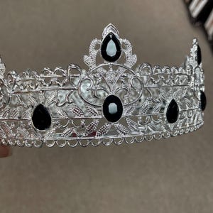 May include: Silver-toned crown with intricate filigree and leaf patterns. The crown is adorned with several large, black teardrop-shaped and oval-shaped gemstones. The design is symmetrical, with a repeating pattern of leaves and curves.