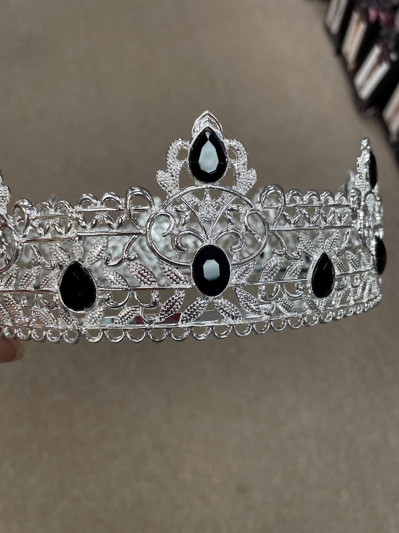 May include: Silver-toned crown with intricate filigree and leaf patterns. The crown is adorned with several large, black teardrop-shaped and oval-shaped gemstones. The design is symmetrical, with a repeating pattern of leaves and curves.
