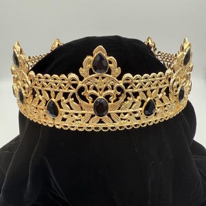 May include: A gold-colored crown with intricate filigree and black gemstone accents. The crown features a series of teardrop-shaped gems and floral designs. The crown is designed for special occasions and events.