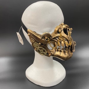 Peut inclure: Un masque de style steampunk dor&eacute; avec des engrenages et une cha&icirc;ne. Le masque est sur une t&ecirc;te de mannequin blanche.