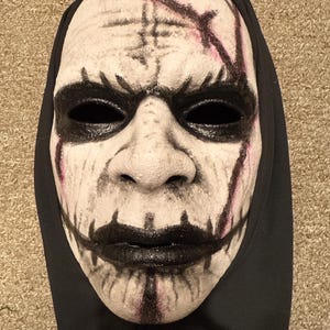 Puede incluir: Una máscara blanca con cuencas de ojos, boca y acentos negros. La máscara tiene una capucha y está pintada con líneas rojas y negras. La máscara tiene una apariencia espeluznante.