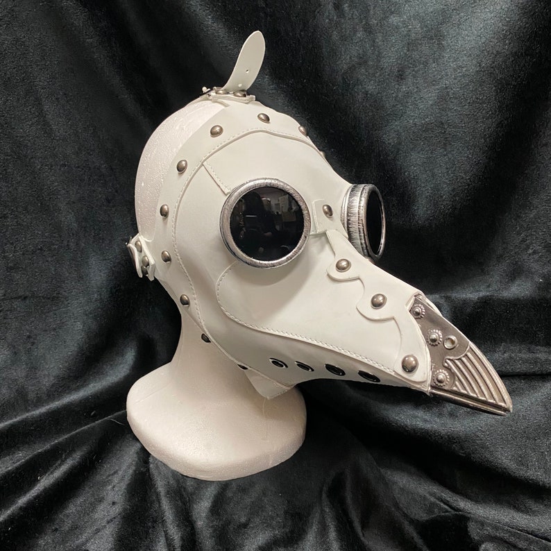 Peut inclure: Masque de m&eacute;decin de la peste en cuir blanc avec des accents argent&eacute;s et deux pi&egrave;ces oculaires rondes noires. Le masque a un long bec pointu.