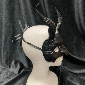 May include: Black masquerade mask with horns and lace detail. The mask is on a white mannequin head.