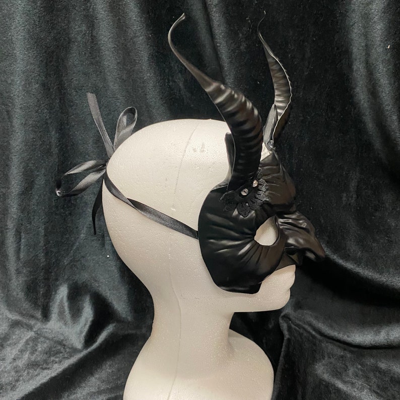 May include: Black masquerade mask with horns and lace detail. The mask is on a white mannequin head.