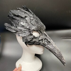 Puede incluir: Una m&aacute;scara de p&aacute;jaro negra y gris con un pico largo y plumas detalladas. La m&aacute;scara est&aacute; hecha de un material texturizado y est&aacute; montada en una cabeza de maniqu&iacute; blanca.