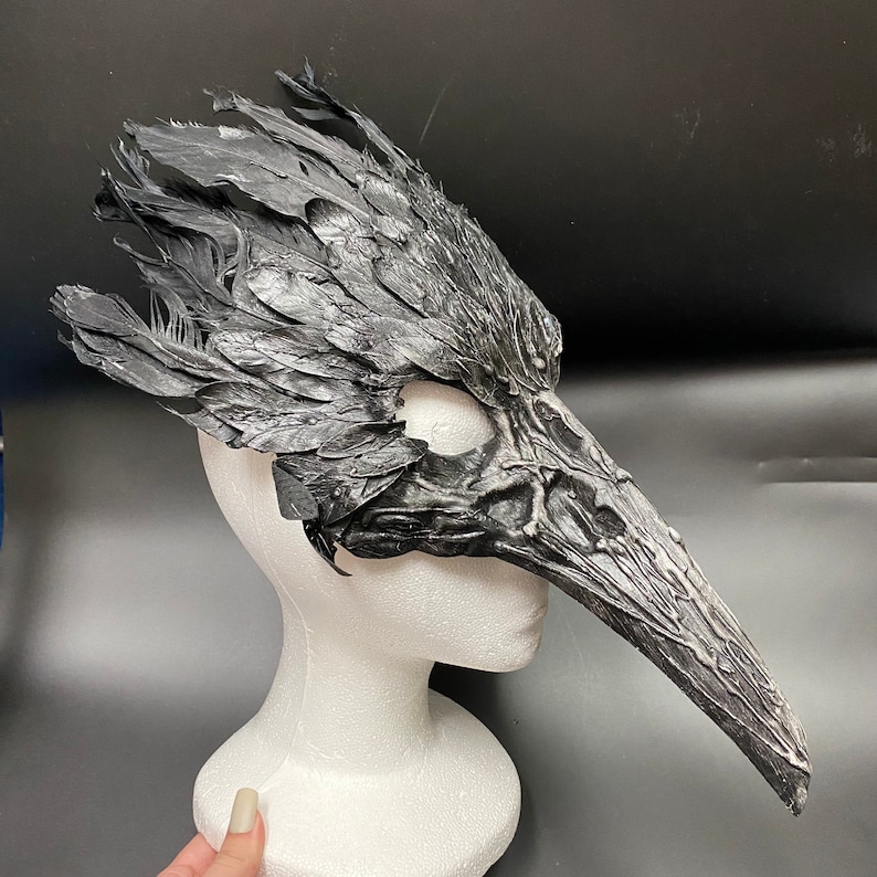 Puede incluir: Una m&aacute;scara de p&aacute;jaro negra y gris con un pico largo y plumas detalladas. La m&aacute;scara est&aacute; hecha de un material texturizado y est&aacute; montada en una cabeza de maniqu&iacute; blanca.