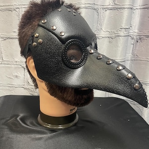 Puede incluir: Una máscara de médico de la peste de cuero negro con un pico largo y detalles metálicos. La máscara tiene una protección para los ojos de malla y se lleva en una cabeza de maniquí.