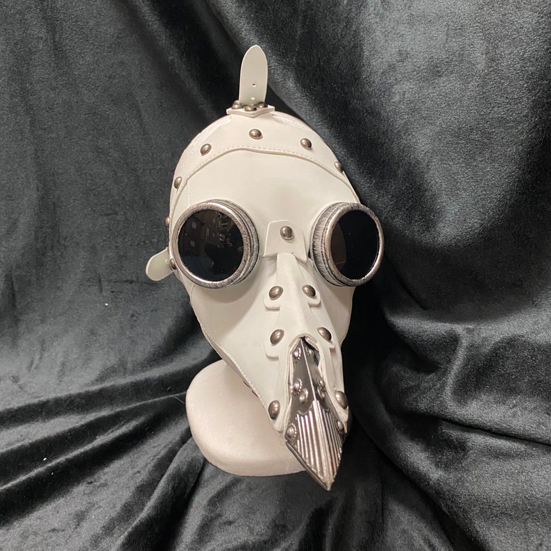 Peut inclure: Masque steampunk en cuir blanc avec des accents argent&eacute;s et deux lunettes rondes noires. Le masque a un long nez en forme de bec.
