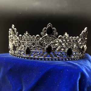May include: A silver-toned crown with intricate filigree detailing and teardrop-shaped black gemstones. The crown sits on a blue velvet surface, showcasing its ornate design. This regal headpiece is suitable for special occasions.