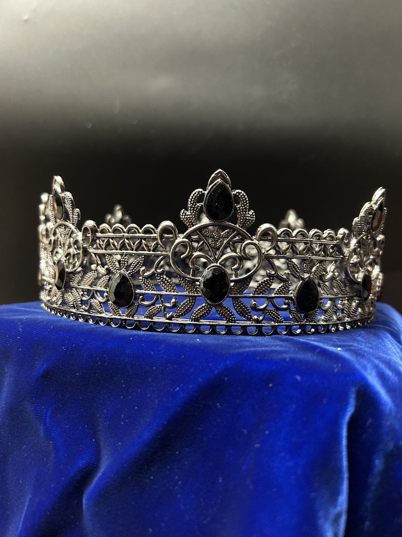 May include: A silver-toned crown with intricate filigree detailing and teardrop-shaped black gemstones. The crown sits on a blue velvet surface, showcasing its ornate design. This regal headpiece is suitable for special occasions.