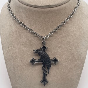May include: A silver chain necklace with a pendant featuring a black raven perched on a silver cross. The cross has a banner with the word "Mort" inscribed on it.