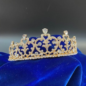May include: A gold tiara with clear rhinestones, featuring a delicate, ornate design. The tiara is on a blue velvet background.