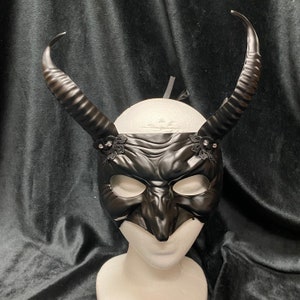 May include: A black masquerade mask with horns and lace accents. The mask has a textured, demonic face design.