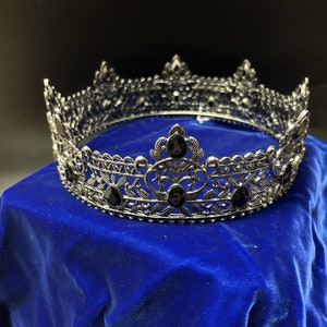 May include: A silver-colored crown with intricate filigree detailing and black gemstone accents. The crown is circular and sits on a blue velvet surface, with a dark background. The crown is likely for a costume or special occasion.