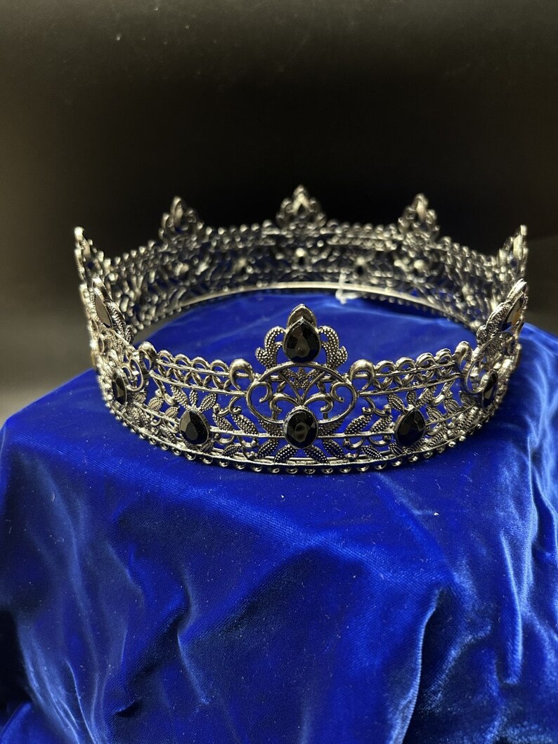 May include: A silver-colored crown with intricate filigree detailing and black gemstone accents. The crown is circular and sits on a blue velvet surface, with a dark background. The crown is likely for a costume or special occasion.