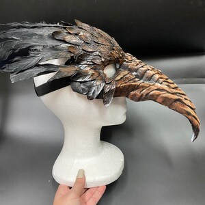 Puede incluir: Una m&aacute;scara de plumas marrones y negras con un pico largo y curvo. La m&aacute;scara est&aacute; unida a una cabeza de poliestireno blanco.