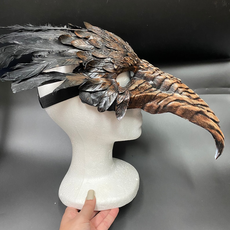 Puede incluir: Una m&aacute;scara de plumas marrones y negras con un pico largo y curvo. La m&aacute;scara est&aacute; unida a una cabeza de poliestireno blanco.
