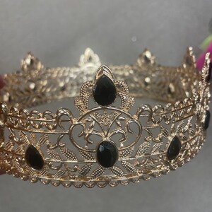May include: A gold-colored crown with intricate filigree detailing and teardrop-shaped black gemstones. The crown features a circular design with decorative floral and leaf patterns, suitable for special occasions or costume use.