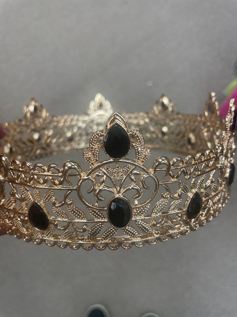 May include: A gold-colored crown with intricate filigree detailing and teardrop-shaped black gemstones. The crown features a circular design with decorative floral and leaf patterns, suitable for special occasions or costume use.