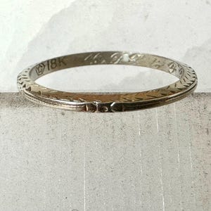 May include: A vintage 18K gold ring with an etched design. The ring has a narrow band with a repeating geometric pattern around the edges. The ring is a light gold color and appears to be well-worn.