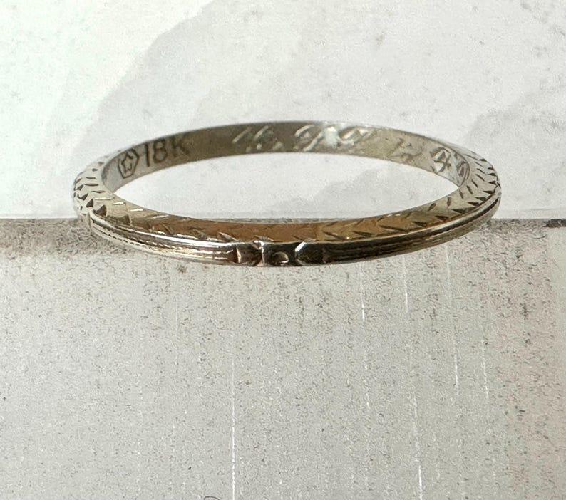 May include: A vintage 18K gold ring with an etched design. The ring has a narrow band with a repeating geometric pattern around the edges. The ring is a light gold color and appears to be well-worn.
