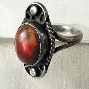 May include: A silver ring featuring an oval-shaped, multi-colored stone with prominent red, orange, and yellow hues. The stone is set in a silver bezel surrounded by a twisted rope border and decorative silver accents at the top and bottom.