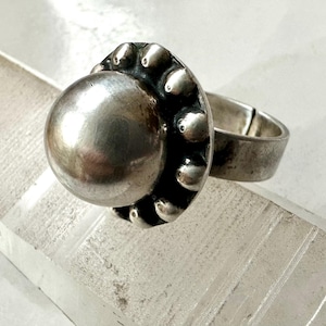 May include: A silver ring with a large, spherical central feature. The ring has a design with small, rounded accents around the central sphere. The band is a simple, polished silver.