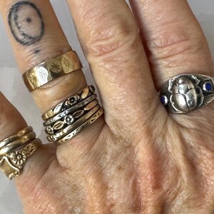 May include: A hand displaying several rings of various designs and materials. The rings include a gold band with starburst engravings, a stack of mixed metal bands, and a silver ring with a scarab beetle design and blue accents. The rings are of different sizes and styles.