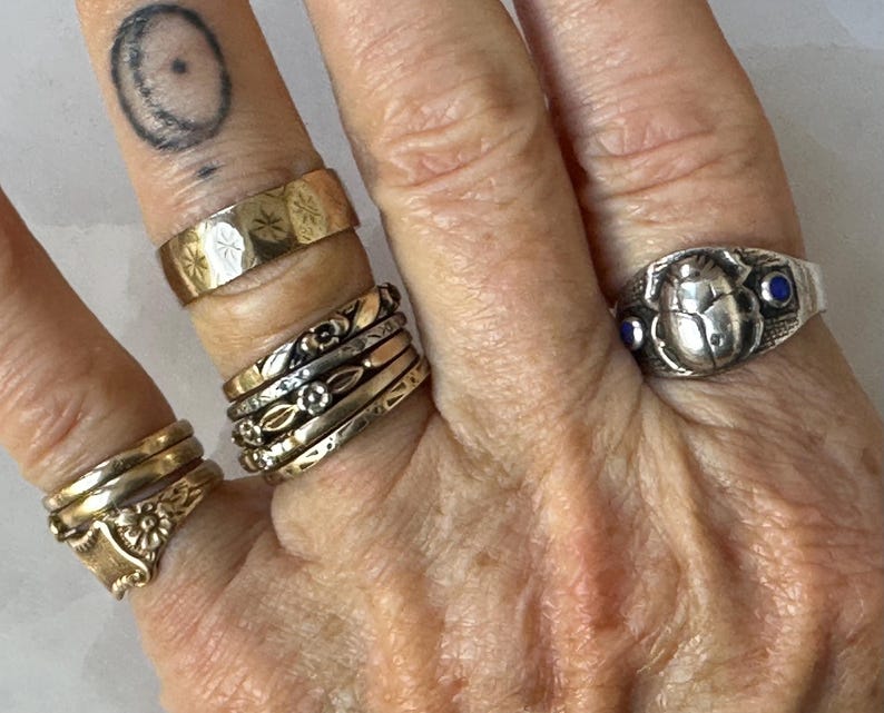 May include: A hand displaying several rings of various designs and materials. The rings include a gold band with starburst engravings, a stack of mixed metal bands, and a silver ring with a scarab beetle design and blue accents. The rings are of different sizes and styles.