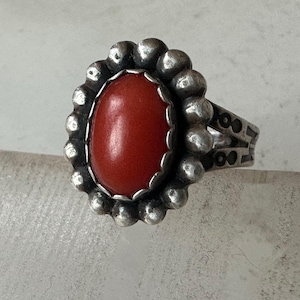 May include: A silver ring with an oval, red gemstone center. The gemstone is surrounded by a beaded silver border. The ring band has a double-band design with decorative cutouts. The ring is displayed on a neutral surface.