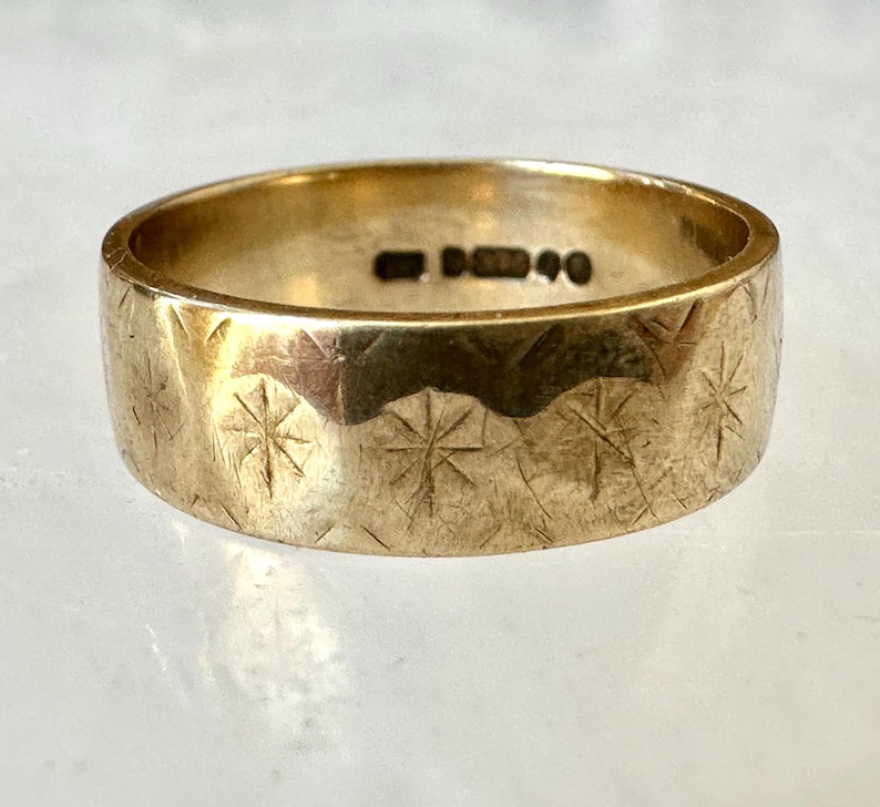 May include: A gold-colored ring with a wide band. The ring's surface is textured with a repeating starburst pattern. The ring has a hallmark stamp on the inside. A piece of jewelry.