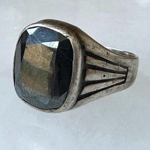 May include: A silver ring with a large, dark, faceted gemstone. The ring has a rectangular face with a slightly rounded top. The band features a geometric design on the sides.