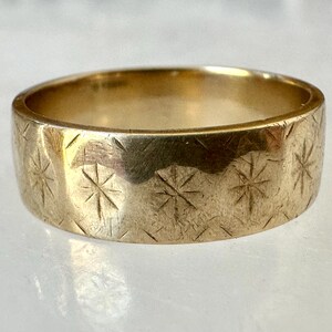 May include: A gold-colored ring with a wide band. The ring's surface is etched with a repeating pattern of starburst designs. The ring is displayed on a light-colored surface.