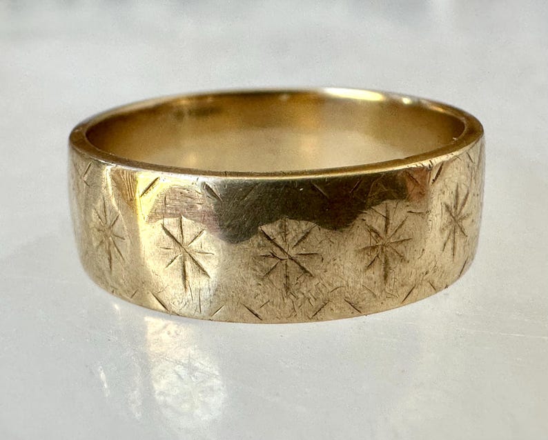 May include: A gold-colored ring with a wide band. The ring's surface is etched with a repeating pattern of starburst designs. The ring is displayed on a light-colored surface.