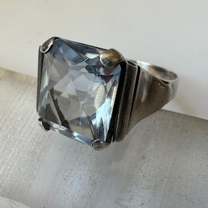 May include: A silver ring featuring a large, rectangular, light blue gemstone. The ring has a thick band and is set with four small, rounded prongs. The gemstone is faceted, catching the light and creating a sparkling effect.