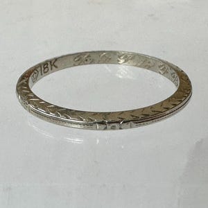 May include: A close-up of a white gold ring with an engraved floral pattern. The ring is marked "18K" and features a delicate design. The ring is a classic piece of jewelry.
