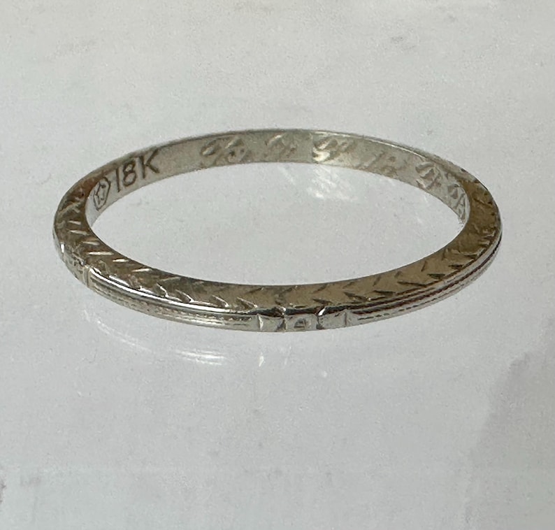 May include: A close-up of a white gold ring with an engraved floral pattern. The ring is marked "18K" and features a delicate design. The ring is a classic piece of jewelry.