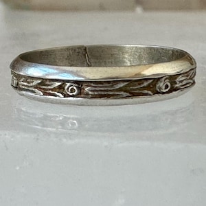 May include: A silver ring with a detailed band featuring a repeating pattern of stylized leaves and circular accents. The ring has a slightly worn appearance, suggesting it is vintage or antique.