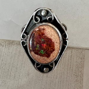 May include: A silver ring featuring a central oval-shaped stone with a textured beige surface and a deep reddish-brown center containing iridescent green and red flecks. The metal setting has a diamond-shaped frame with decorative scrollwork.