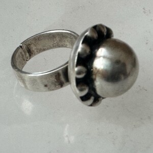 May include: A silver ring featuring a large, polished, spherical centre stone. The adjustable band shows signs of wear. The setting around the stone has small, decorative beads. The ring is likely made of metal.