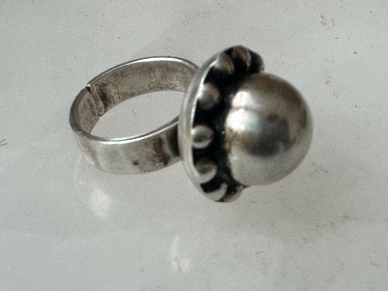 May include: A silver ring featuring a large, polished, spherical centre stone. The adjustable band shows signs of wear. The setting around the stone has small, decorative beads. The ring is likely made of metal.