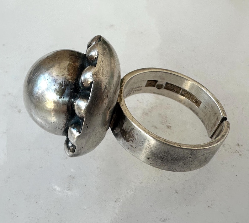May include: A silver ring with a large, spherical design. The ring features a textured, orb-like centerpiece with smaller, rounded accents. The band is thick and has a worn appearance, suggesting an antique or vintage style.