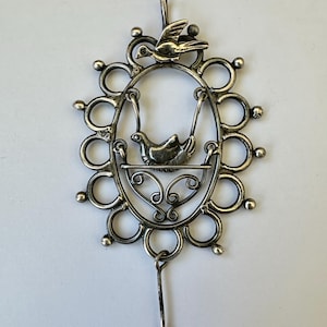 May include: A silver-toned pendant featuring a bird in a swing within an oval frame, surrounded by decorative circles and a small bird at the top. The pendant has a loop at the top for hanging and a dangling element at the bottom. The design is intricate and detailed.